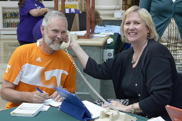Woman holding up a phone to a man's ear while he writes a check for charity.