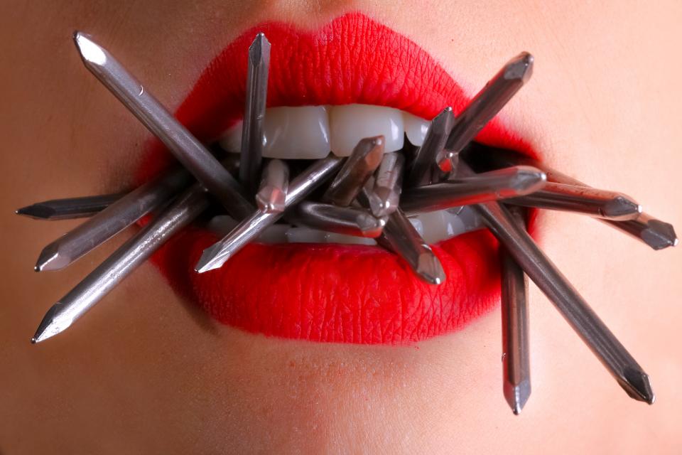 A close up of a woman's lips in bright lipstick with nails coming out of her mouth.