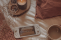 A Nintendo Switch lite on a bed next to a mug of tea, a burnt orange pillow and blanket, and a lit candle