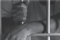 A fair skinned person in a white t-shirt stands within a jail cell with white painted bars. He drapes on wrist over the lock and grips a bar with his other hand.